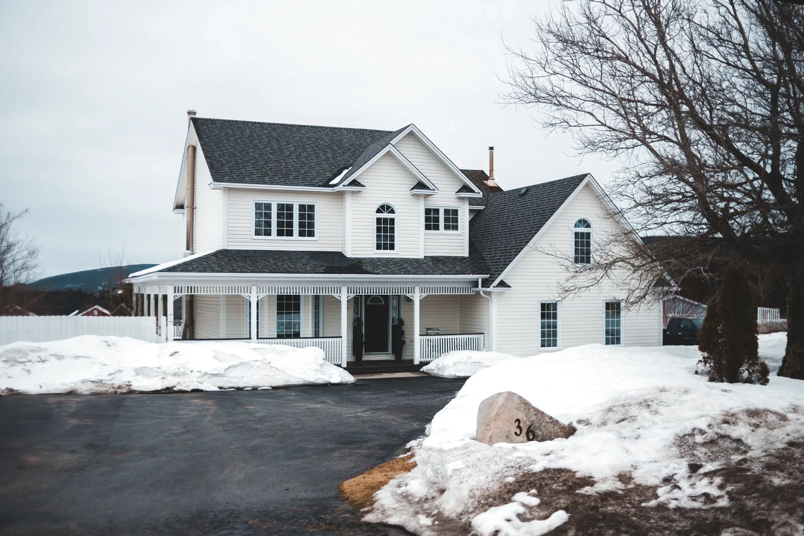A white family home with a wraparound porch and snow across the yard.